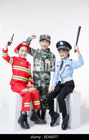 Three children dressed in different professions, sitting on a white bench, smiling and playing with their props. Stock Photo
