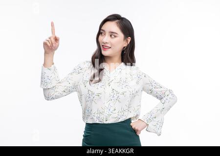 A young woman with a bright smile and raised index finger points to something above her, standing against a plain white background. Stock Photo