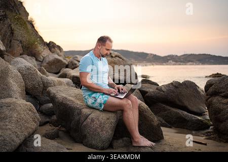 Man working remotely on laptop at rocky Greece beach Stock Photo