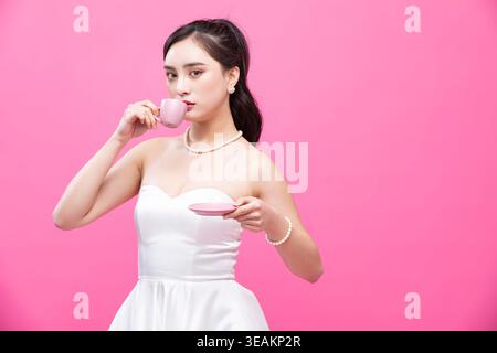 A young woman in a white wedding dress sips tea from a delicate pink cup in a serene, elegant setting. Stock Photo