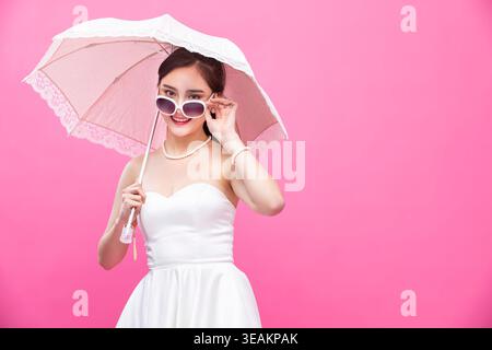 A young woman in a white wedding dress holds a parasol and smiles, standing in front of a bright pink background. Stock Photo