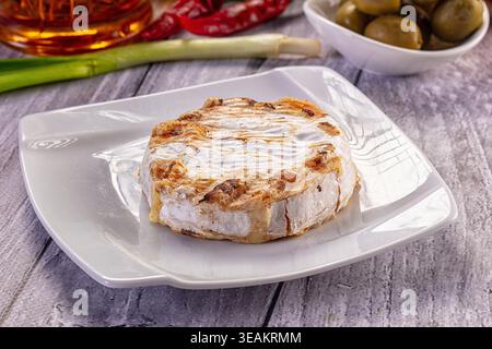 A wheel of golden-brown grilled Camembert cheese served on a modern white plate. The rustic scene includes olives, peppers, and green onion on a gray Stock Photo