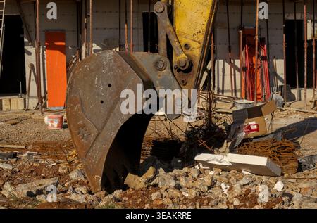 Yellow excavator bucket close-up at a construction site with building scaffolding and formwork in the background. Stock Photo
