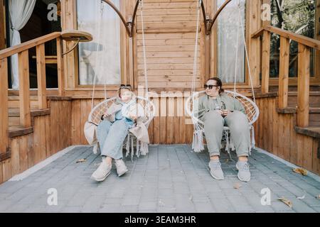 Two Women Relaxing in Hanging Chairs on a Wooden Porch Stock Photo