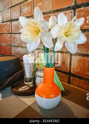 Riga, Latvia - April 22, 2026:Fresh white daffodils in a small vase on a cozy cafe table against a rustic brick wall. Warm tones and simple decor crea Stock Photo