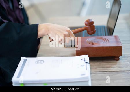 A judge's hand is holding a gavel, sitting at a desk with a laptop and a stack of papers. Stock Photo