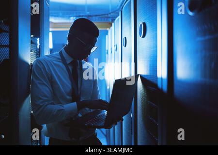 IT, man or typing on laptop in server room for cybersecurity, software upgrade or diagnostics. Technician, black person or tech in dark data center Stock Photo
