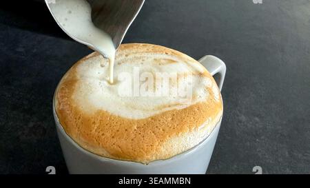 Close-up of steaming milk being poured into a gray coffee cup, creating a frothy layer on top of the espresso, with a dark surface in the background Stock Photo