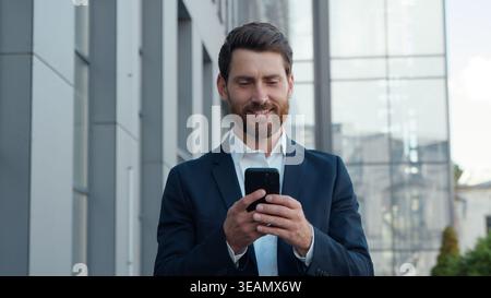 Smiling happy Caucasian business man walking on street with mobile phone middle-aged businessman with smartphone walk going in city leaving office Stock Photo