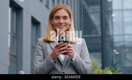 Adult Caucasian business woman using mobile phone smiling looking at camera cheerful businesswoman female with smartphone marketing manager scrolling Stock Photo