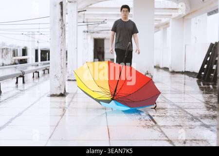 Young man standing in wet train station with colorful umbrella lying on floor, urban rainy day scene Stock Photo