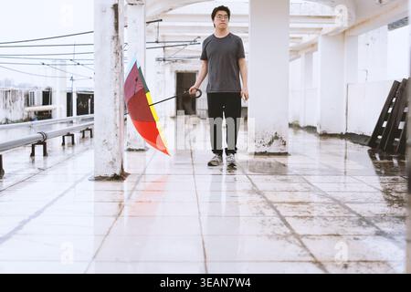 Young man walking with colorful umbrella in wet urban corridor, modern architecture, reflective Stock Photo