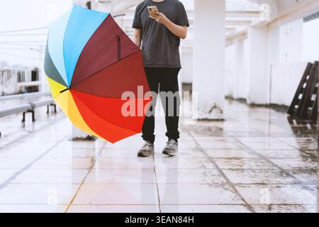 Man holding colorful rainbow umbrella while using smartphone on wet urban walkway Stock Photo