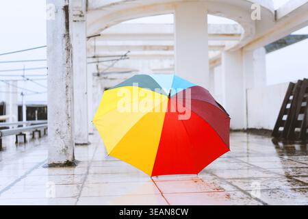 Colorful rainbow umbrella resting on wet tiled floor under white arches in modern station or plaza Stock Photo