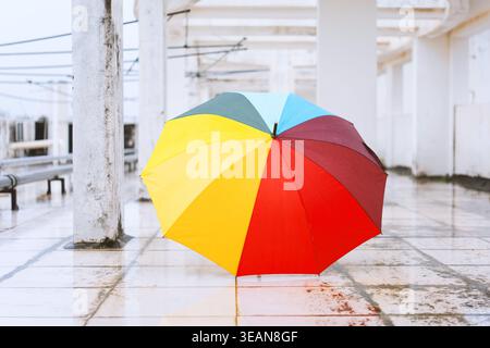 Colorful rainbow umbrella standing alone on wet tiled floor under white concrete structure Stock Photo