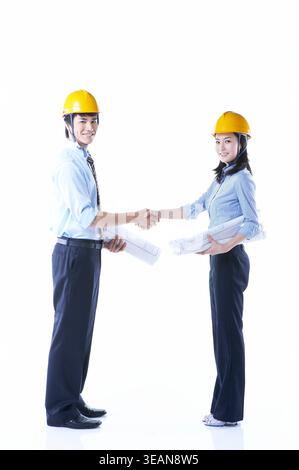 Two engineers in hard hats shake hands while holding blueprints, standing against a white background. Stock Photo