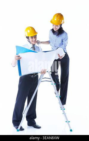Two engineers in hard hats reviewing blueprints on a ladder. Stock Photo