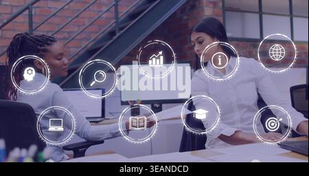 Two coworkers in blue blouse and white shirt reviewing documents in office with monitors and icons. Collaboration, analytics, spreadsheets, data, team Stock Photo