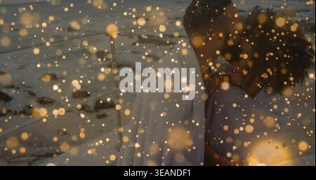 Embracing couple leaning in on dusk shore, wrapped in pale shawl with golden bokeh, copy space. Romance, beach, twilight, intimacy, warm, moody, light Stock Photo