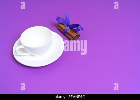 White Cup and Cinnamon Sticks with Purple Ribbon on a Vibrant Purple Background Stock Photo
