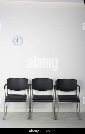 Three empty black chairs are arranged in a row against a white wall, with a clock on the wall above them. Stock Photo