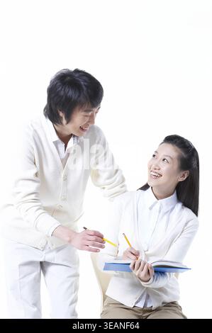 A young Asian man is helping a young Asian woman with her homework in a bright, white classroom. Stock Photo