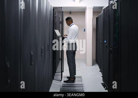 Engineer, black man or typing on laptop in server room for console access, power backup or update. Computer technician, person and inspection in data Stock Photo