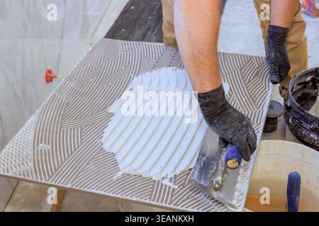 Tiling worker uses trowel to spread adhesive on tile during flooring project in construction area. Stock Photo
