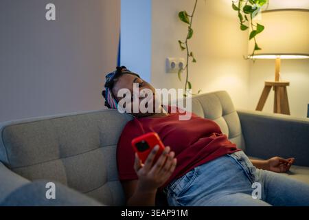Tired black woman leaning back on couch with smartphone feeling drained, bored. Internet addiction Stock Photo