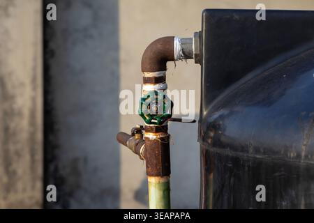 Close-up of a green gate valve attached to a rusty iron pipe on a black plastic water storage tank. Ideal for plumbing services, water management, and Stock Photo