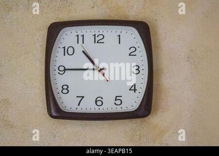 Front view of a classic square wall clock with rounded corners hanging on a beige textured wall. Ideal for time management, office decor, and interior Stock Photo