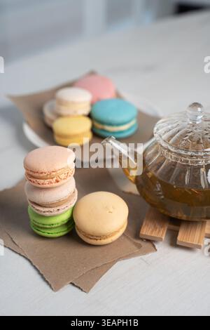 close-up of pastel macarons stacked and scattered beside a clear glass teapot of amber herbal tea on torn brown paper over a light wooden table, soft Stock Photo
