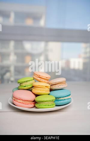 assorted pastel macarons artfully stacked on a white plate by a bright window in a modern apartment, soft-focus urban backdrop visible through glass, Stock Photo