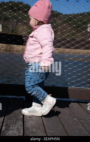 side-profile of a toddler wearing a pink beanie and floral jacket standing on a wooden dock by a lake, blue sky, wire-fence railing, denim jeans and w Stock Photo