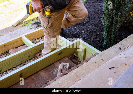 Worker is using drill to secure steps wood pieces while standing on step outside, surrounded by construction materials. Stock Photo