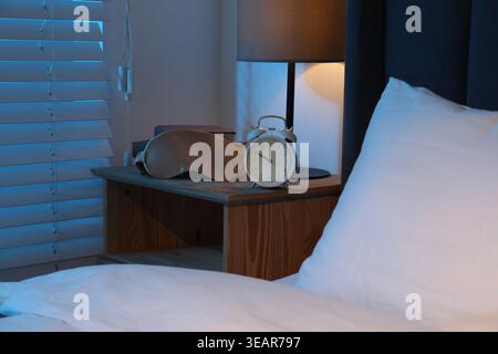 Alarm clock, sleeping mask, books and lamp on bedside table indoors, closeup Stock Photo