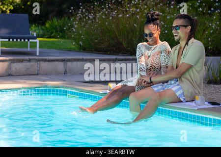 African American couple sitting on towel at pool edge holding hands, lemon glasses, copy space. Romance, leisure, outdoor, backyard, sunlit, relaxed, Stock Photo