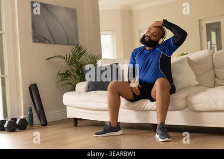 African American adult male sitting cooling down on cream sofa with dumbbells, headphones, water. Couch, cooldown, home, fitness, weights, relaxation, Stock Photo