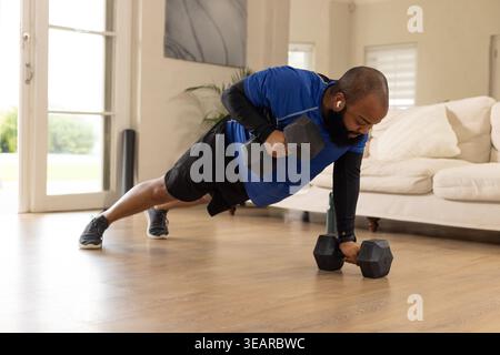 African American man doing renegade row at home wearing workout gear with hex dumbbells and earbuds. Weights, plank, training, fitness, sunlit, apartm Stock Photo