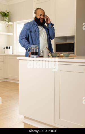 African American man laughing while holding headphone at kitchen island with French press in denim. Coffee, headset, joy, bright, modern, mug, brewing Stock Photo