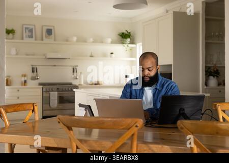African American man working on laptop and monitor at table in kitchen in denim shirt. Homeoffice, naturallight, wood, dualdisplay, cables, greenery, Stock Photo