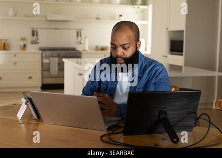 African American man wearing denim shirt sitting at table in kitchen, using laptop, tablet, phone. Workspace, home, modern, minimal, bright, neutral, Stock Photo
