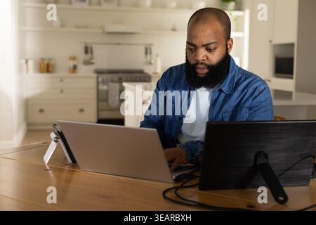 African American man sitting at wooden table in kitchen, working on laptop and tablet. Homeoffice, remotework, multiscreen, cables, interior, denim, b Stock Photo