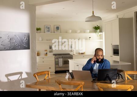 Adult African American man sitting at kitchen table calling on phone, using two laptops, copy space. Multitask, remote, workfromhome, homeoffice, prod Stock Photo