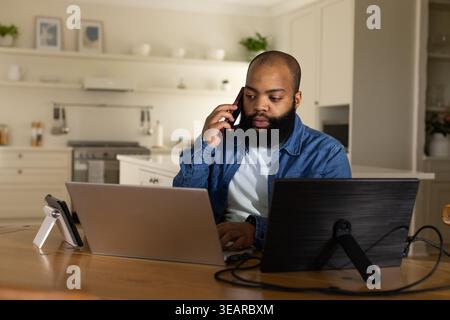 Bearded man working in kitchen using dual screens and speaking on phone stand, in denim jacket. Remote, laptops, cables, plants, modern, homeoffice, p Stock Photo