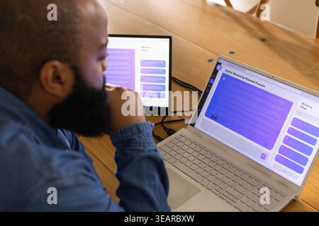 Bearded man sitting at wooden table working on laptop and external monitor with cables, copy space. Workspace, business, modern, advertising, planning Stock Photo