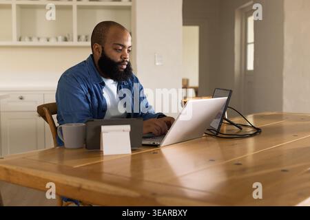 African American man working on laptop at home kitchen table with tablet and mug. Remote, workspace, homeoffice, monitor, minimalist, daylight, wooden Stock Photo