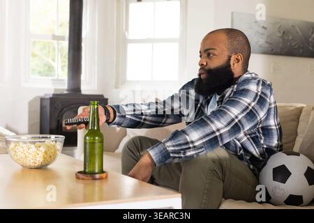 African American adult man leaning reaching for remote watching TV at home with popcorn, copy space. Sofa, television, viewer, daylight, livingroom, s Stock Photo