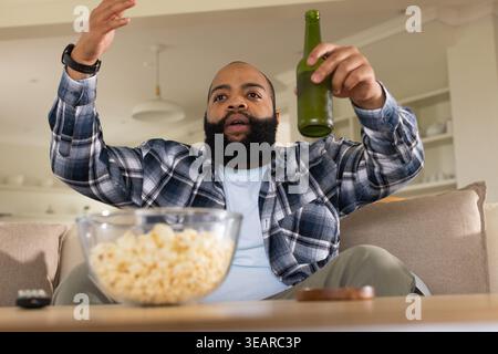 Adult African man sitting on couch in plaid holding green bottle gesturing by popcorn bowl, remote. Beard, casual, lounge, wooden, pendant, lighting, Stock Photo