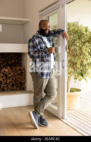 African American man leaning on sliding glass door at home holding white ceramic mug and smiling. Interior, daylight, casual, lifestyle, contemporary, Stock Photo